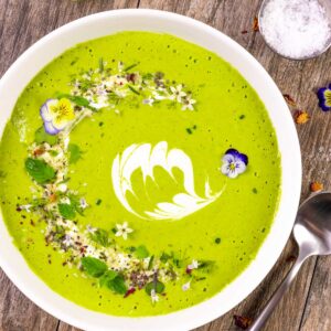 Cucumber gazpacho with avocado in a white bowl with herb seeds, mint, flower petals and a design of sour cream in the center.