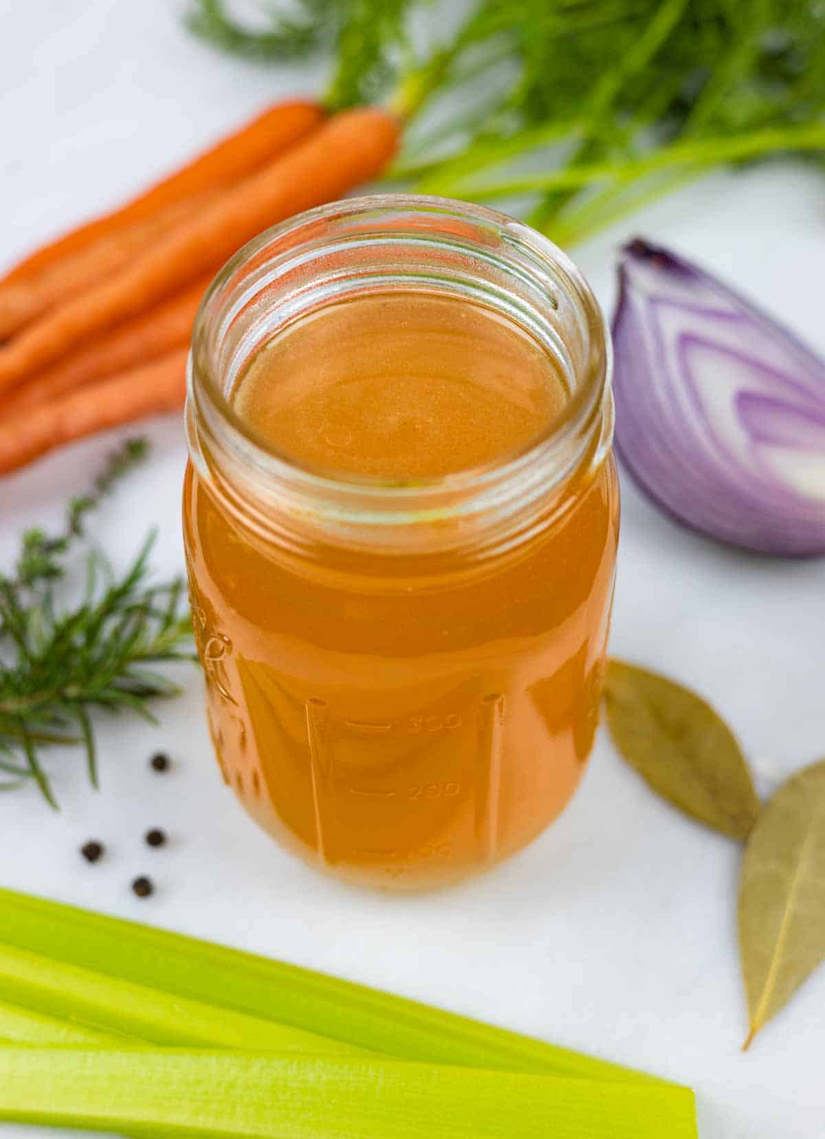 Chicken bone broth in a mason jar on a white board with bunch of carrots, red onion quarter, celery sticks and spices.