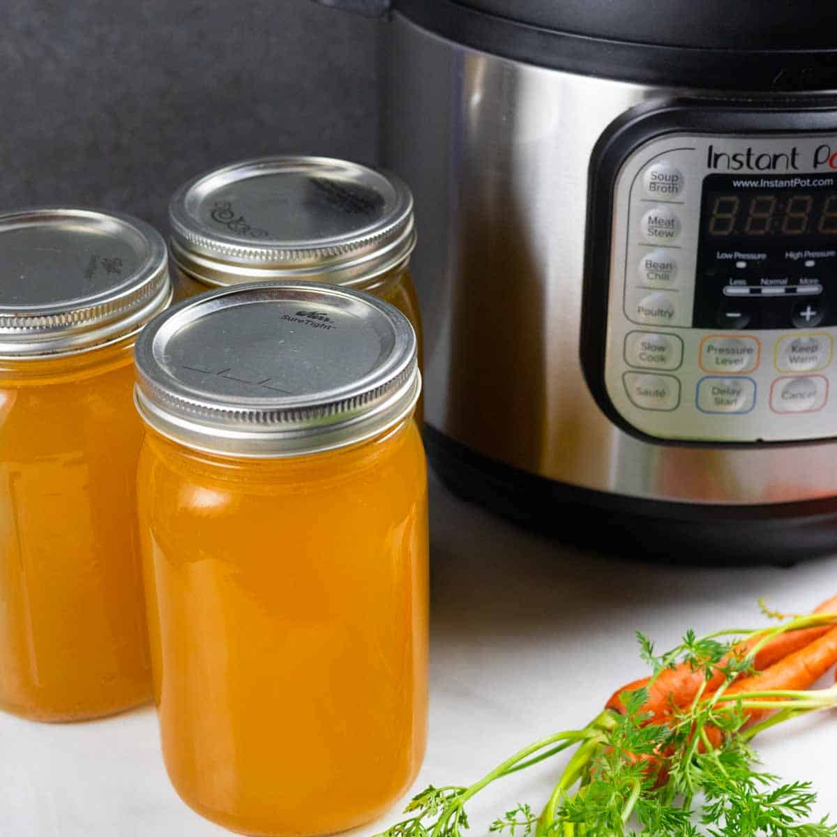 Three lidded quart jars of finished chicken bone broth on a white board, sitting next to an Instant pot with a small bunch of orange carrots in front.