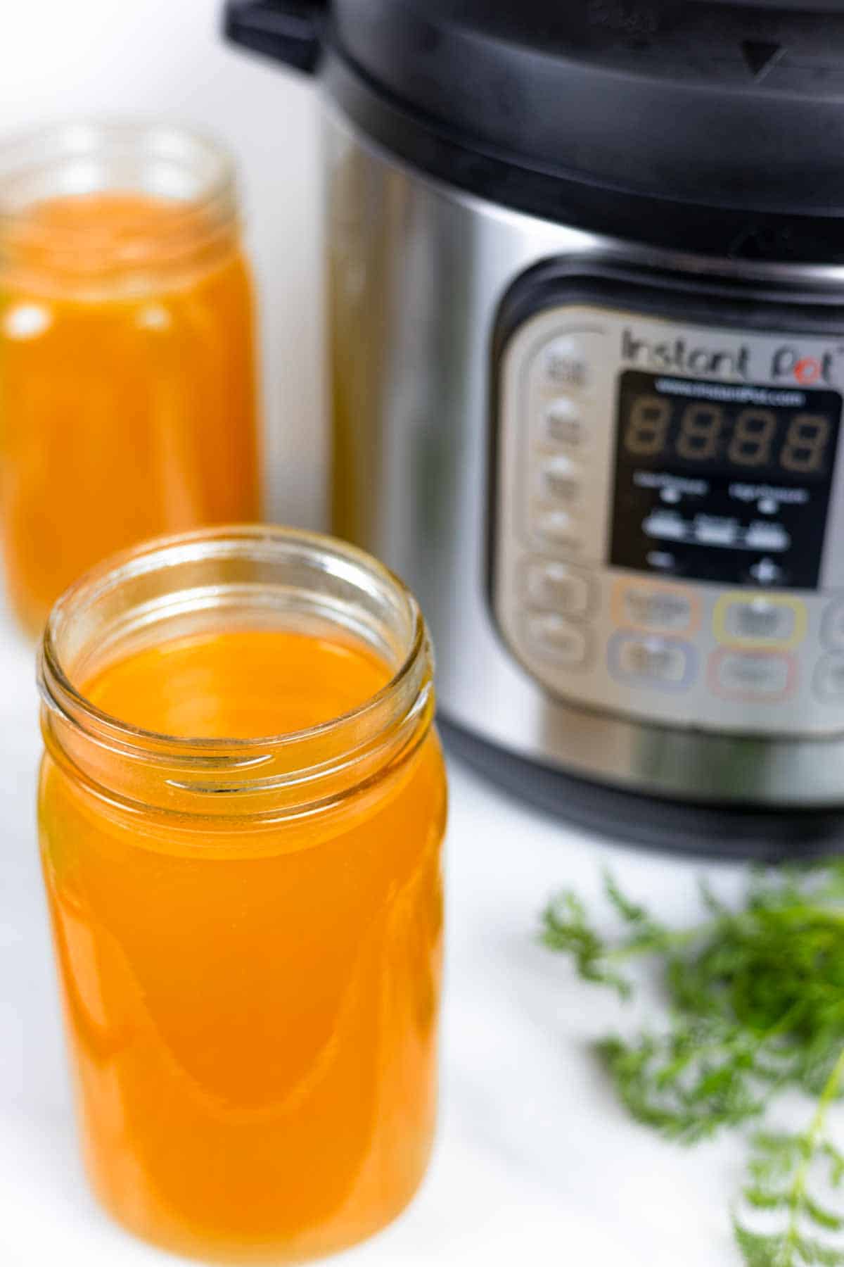 Quart jars of chicken broth on a white board in front of an Instant Pot with herbs sprinkled in front.