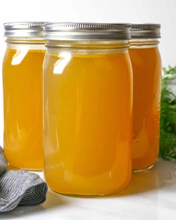 Three quart jars of finished chicken bone broth on a white board with a grey napkin in front and a carrot bunch behind.