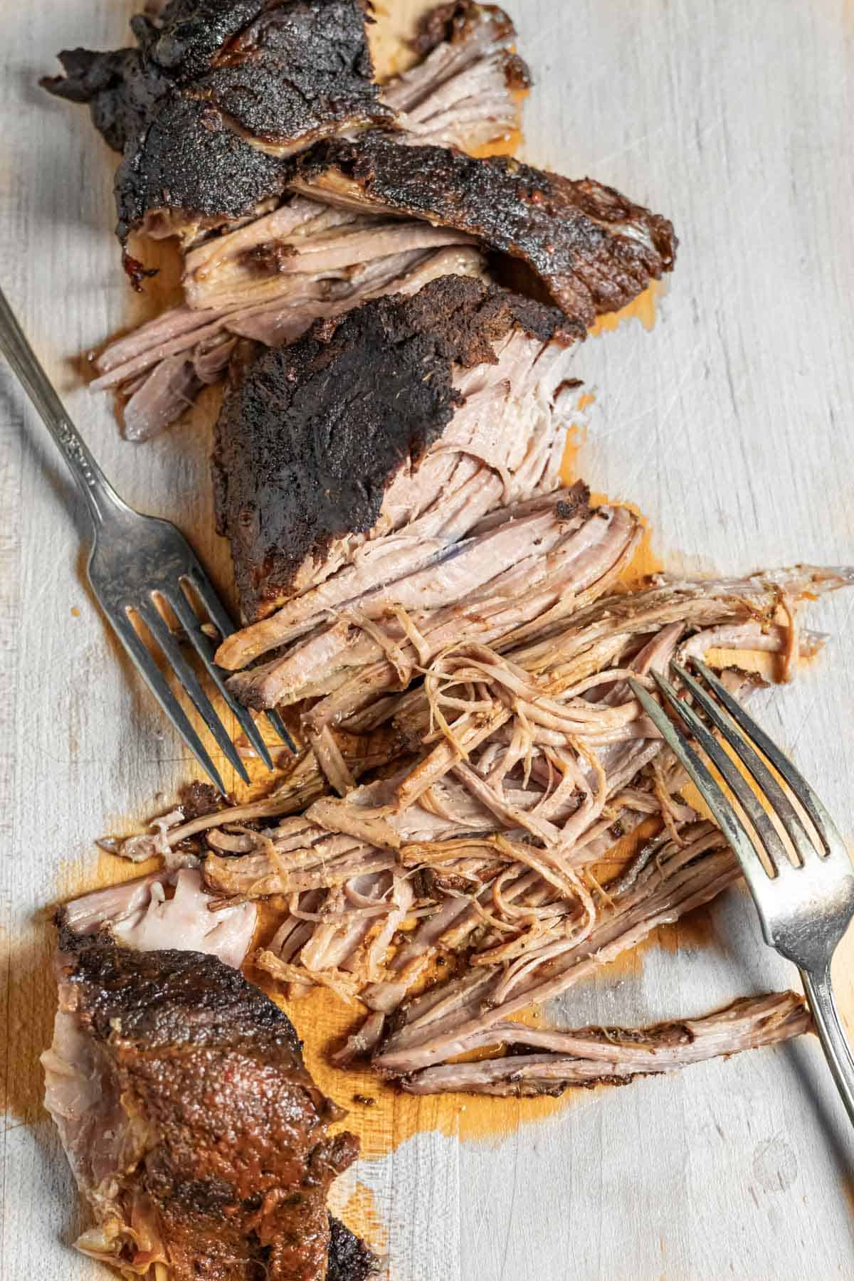 Tender pork partly shredded on a wood board with two forks and showing the crusty bark.