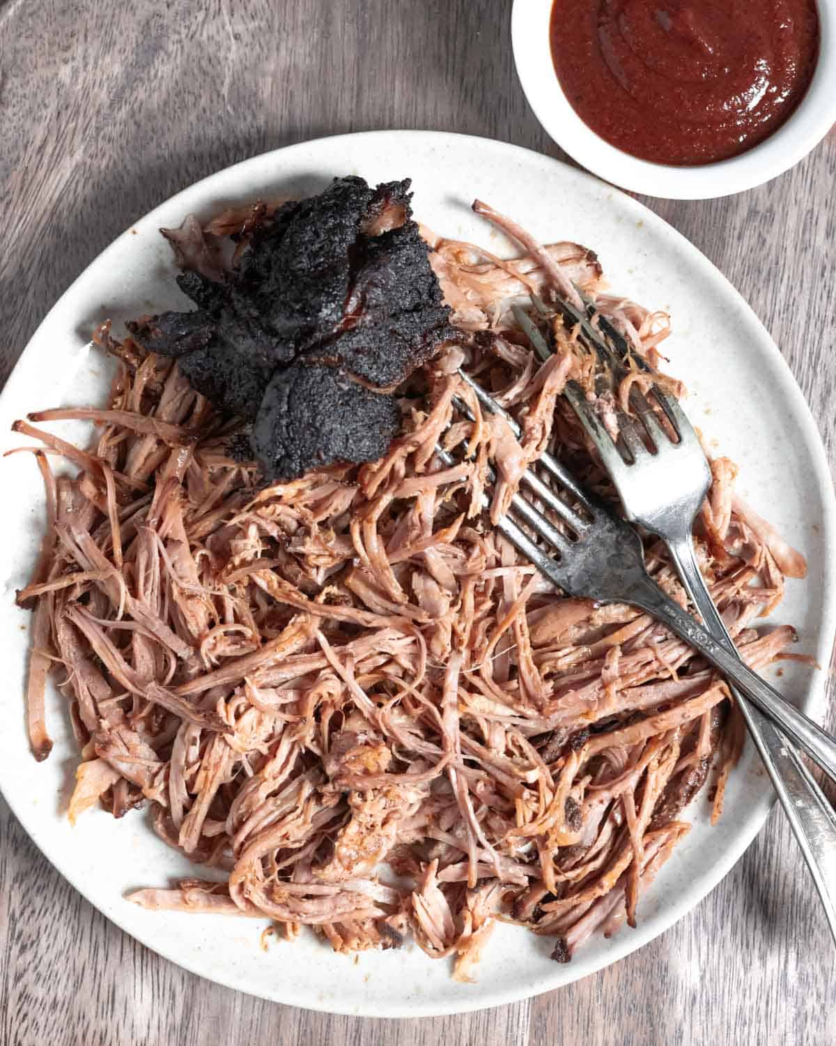 White plate full of shredded pork with a barky chunk, two forks, and a ramekin of sauce.