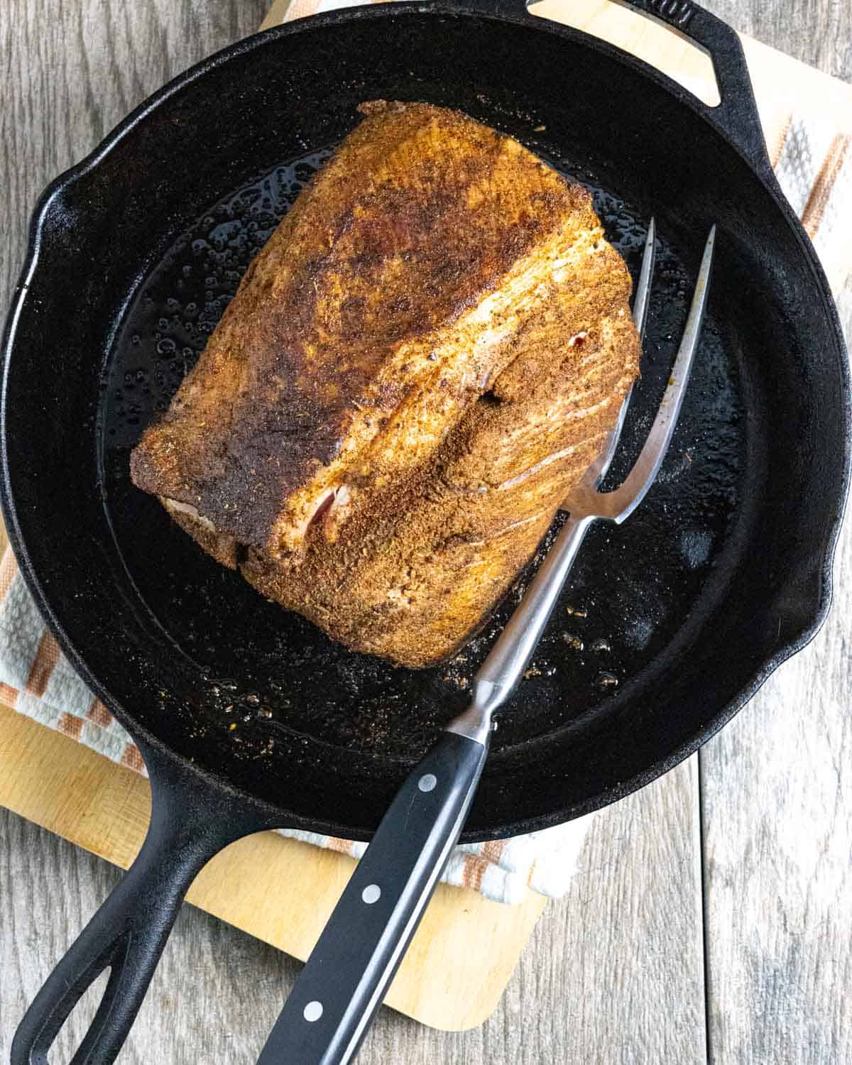 Browned seasoned pork shoulder roast in a cast iron skillet with a large carving fork in the pan.