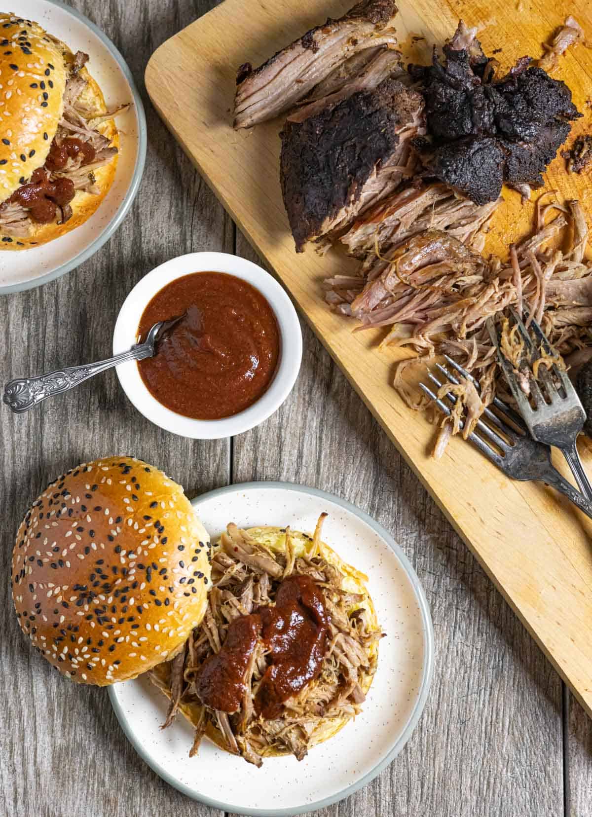 Board of pulled pork with plates of sesame buns filled with pork and sauce with a small bowl of sauce.