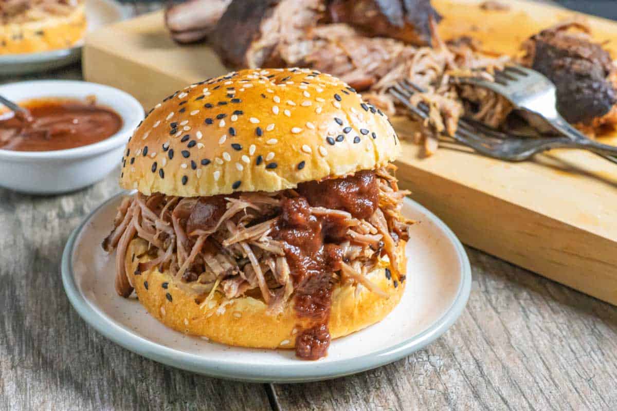 Pulled pork and sauce stacked on a sesame seed bun on a small plate with more pork behind it on a board.