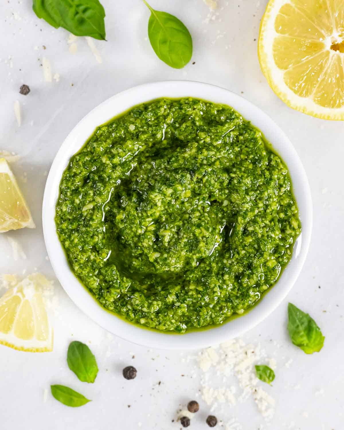 Pesto without pine nuts in a ramekin on a white board with lemon slices, basil leaves and peppercorns.