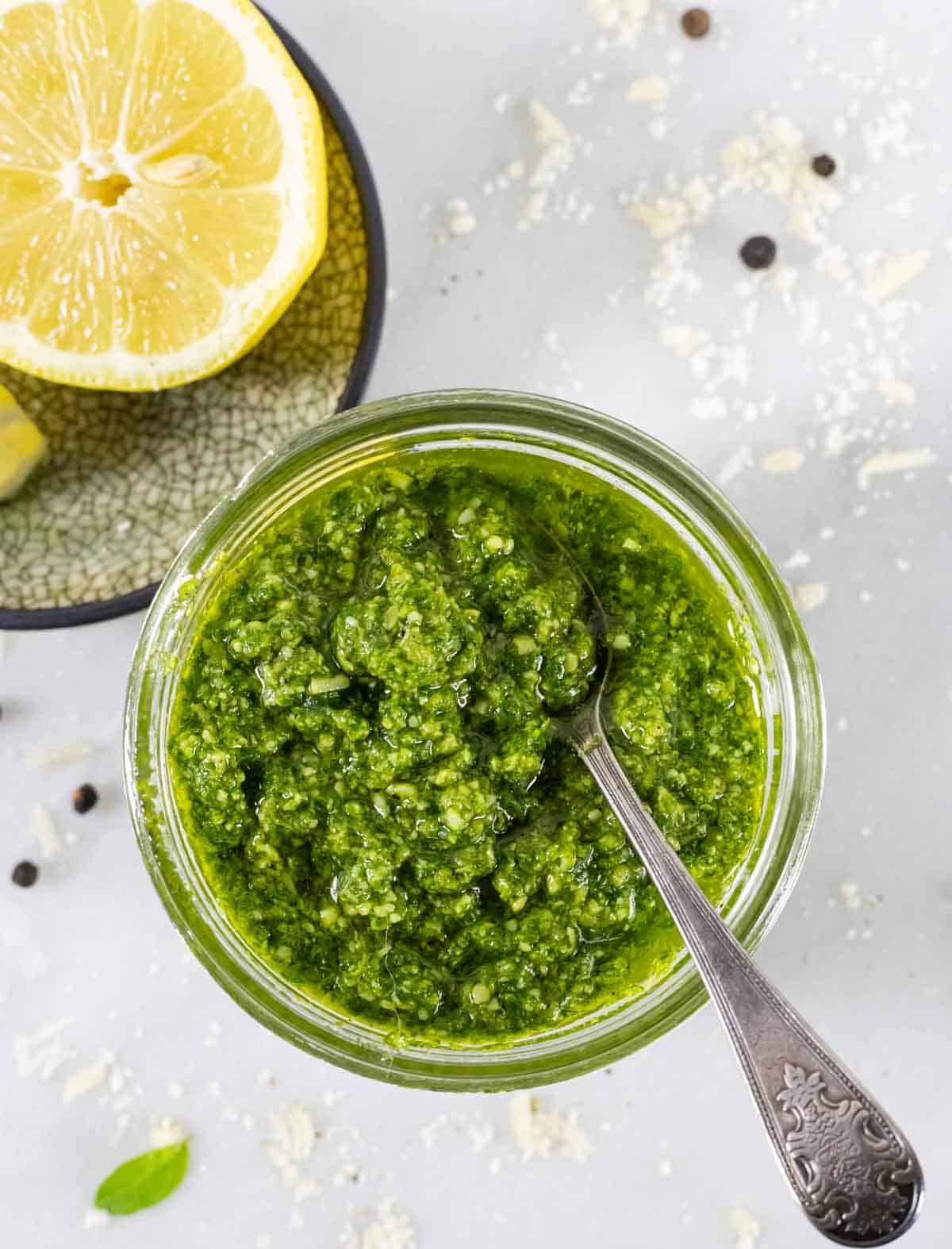 Looking down into jar of pesto with small silver spoon in it with lemon halves on the side.
