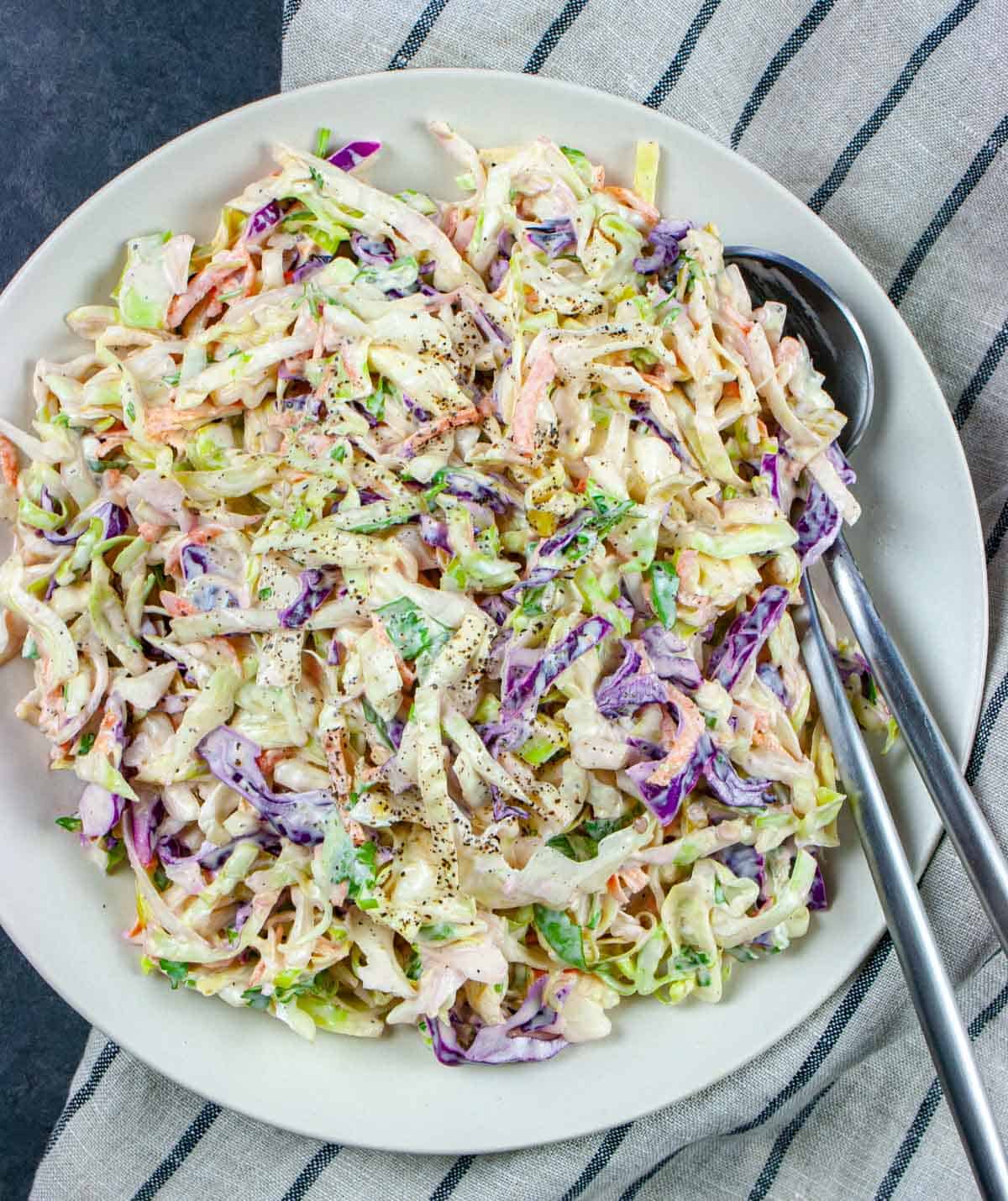 White bowl of slaw sprinkled with black pepper with a fork and spoon.