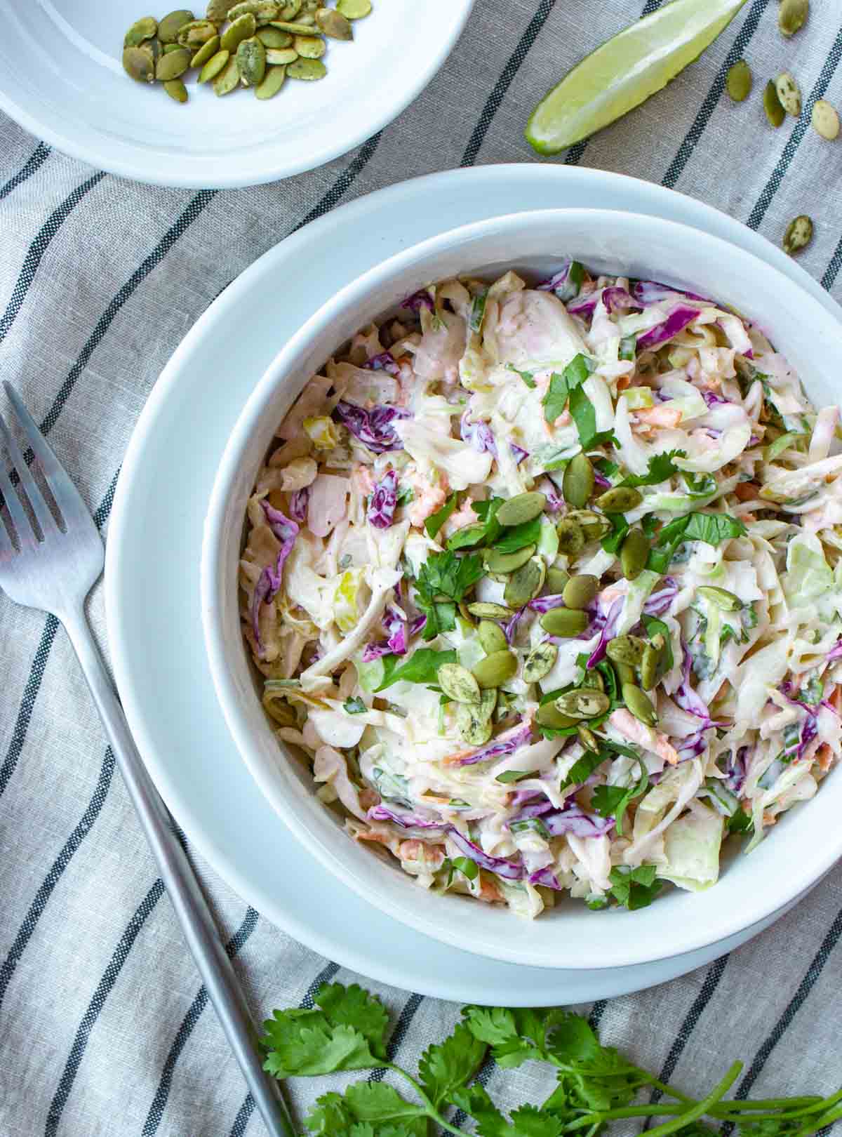 Serving of red and green coleslaw in a white bowl on a white plate with garnish of cilantro, lime wedge and pumpkin seeds.