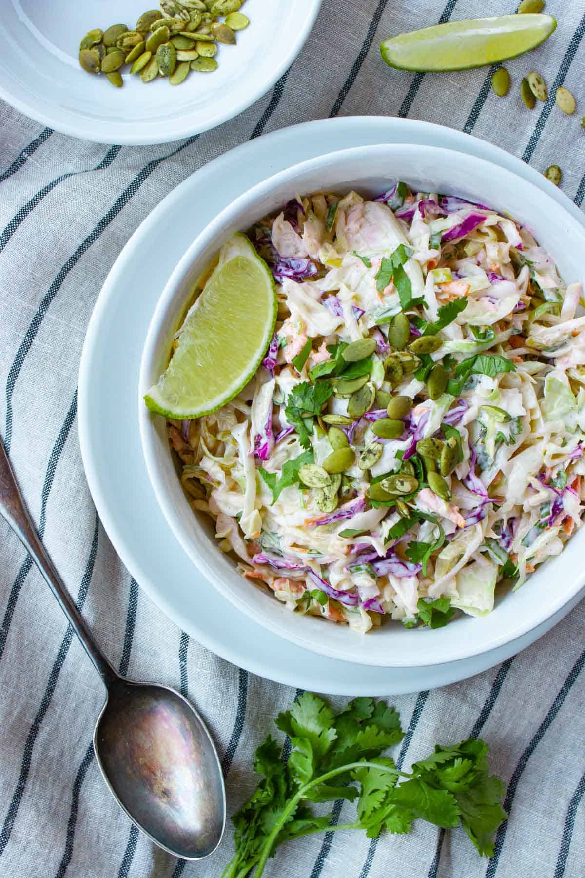 Coleslaw in white bowl with spoon and cilantro on a white and blue napkin.