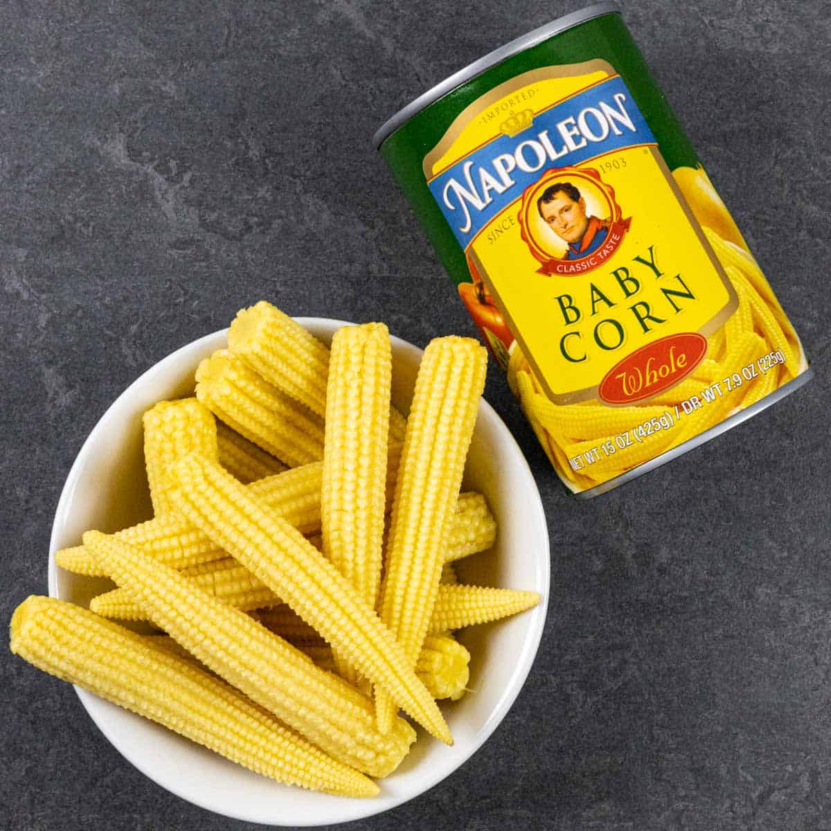 Small white bowl of whole canned baby corn cobs on a grey board next to the can showing its front label.