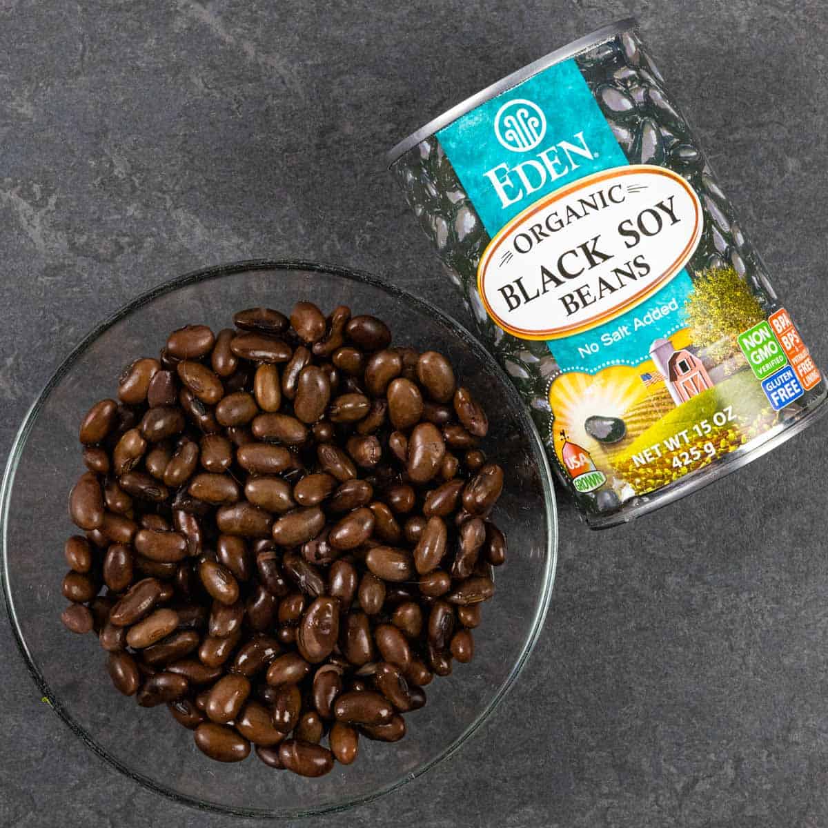 Glass bowl of black soy beans on a grey board next to the can of beans on its side.