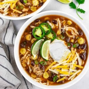 Serving of low carb chicken taco soup in a white bowl topped with lime wedges, sour cream, shredded cheese and sliced jalapeno.