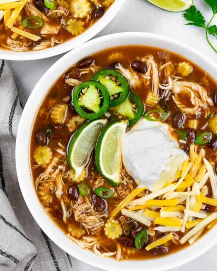 Serving of low carb chicken taco soup in a white bowl topped with lime wedges, sour cream, shredded cheese and sliced jalapeno.