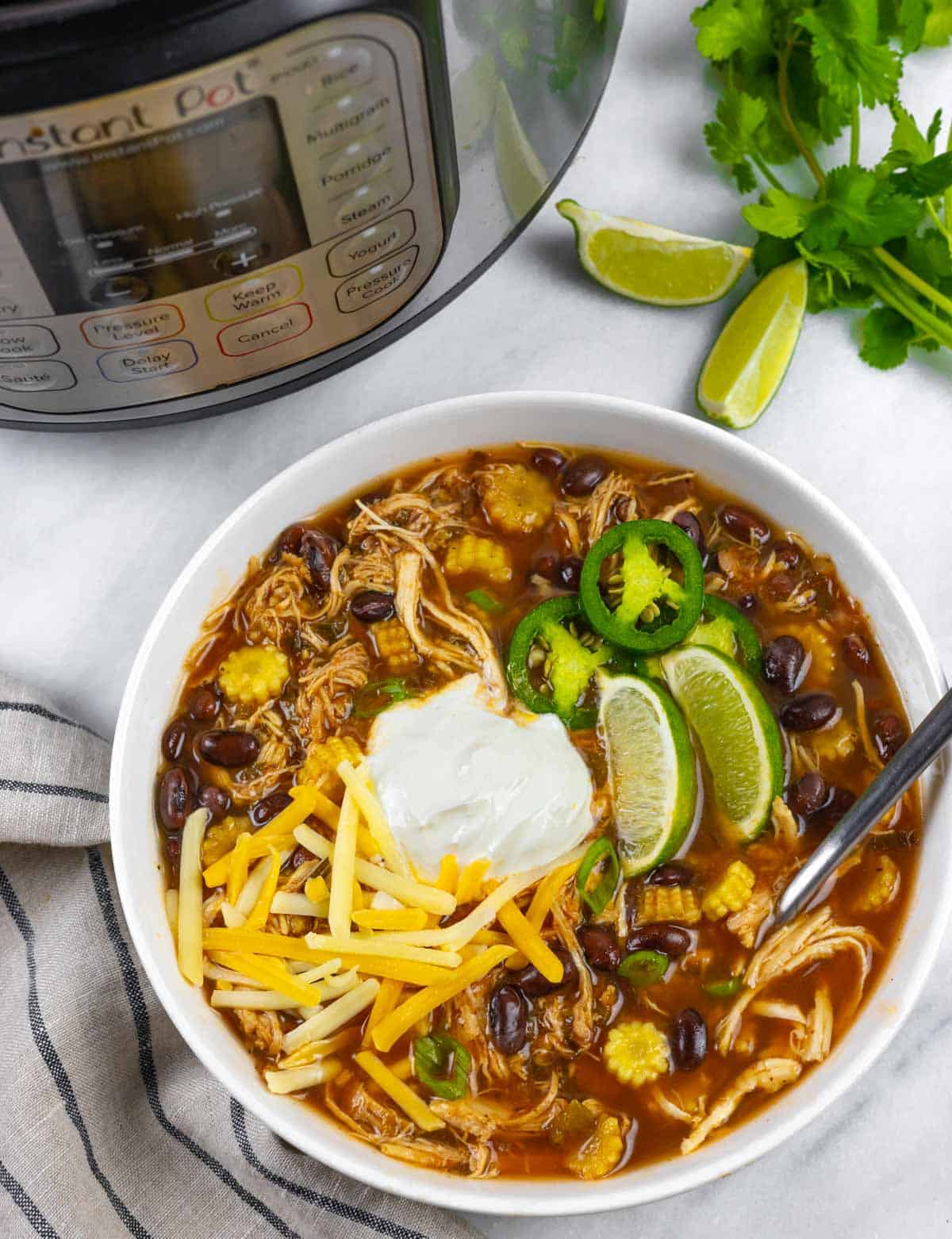 White bowl of chicken taco soup with a spoon, sitting on a marble board next to an instant pot, with lime wedges and cilantro as garnish.