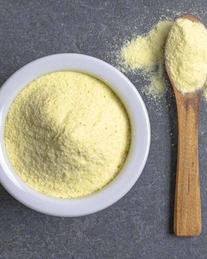 Asafoetida powder piled in a small white bowl with a wood spoon spilling over with spice next to the bowl.