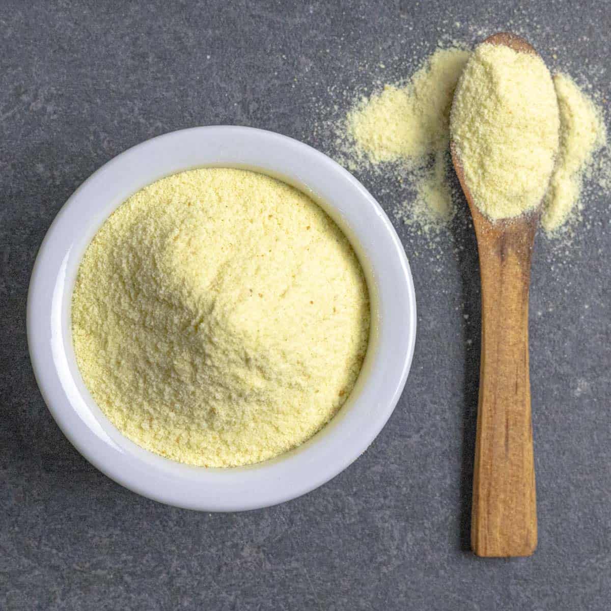 Asafoetida powder piled in a small white bowl with a wood spoon spilling over with spice next to the bowl.