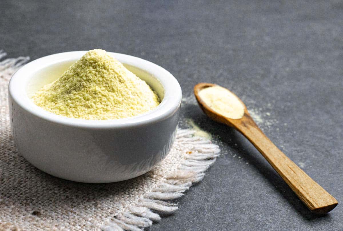 Hing powder spice piled in a white bowl on a cloth placemat with a small wood spoon of spice sitting on a grey board.