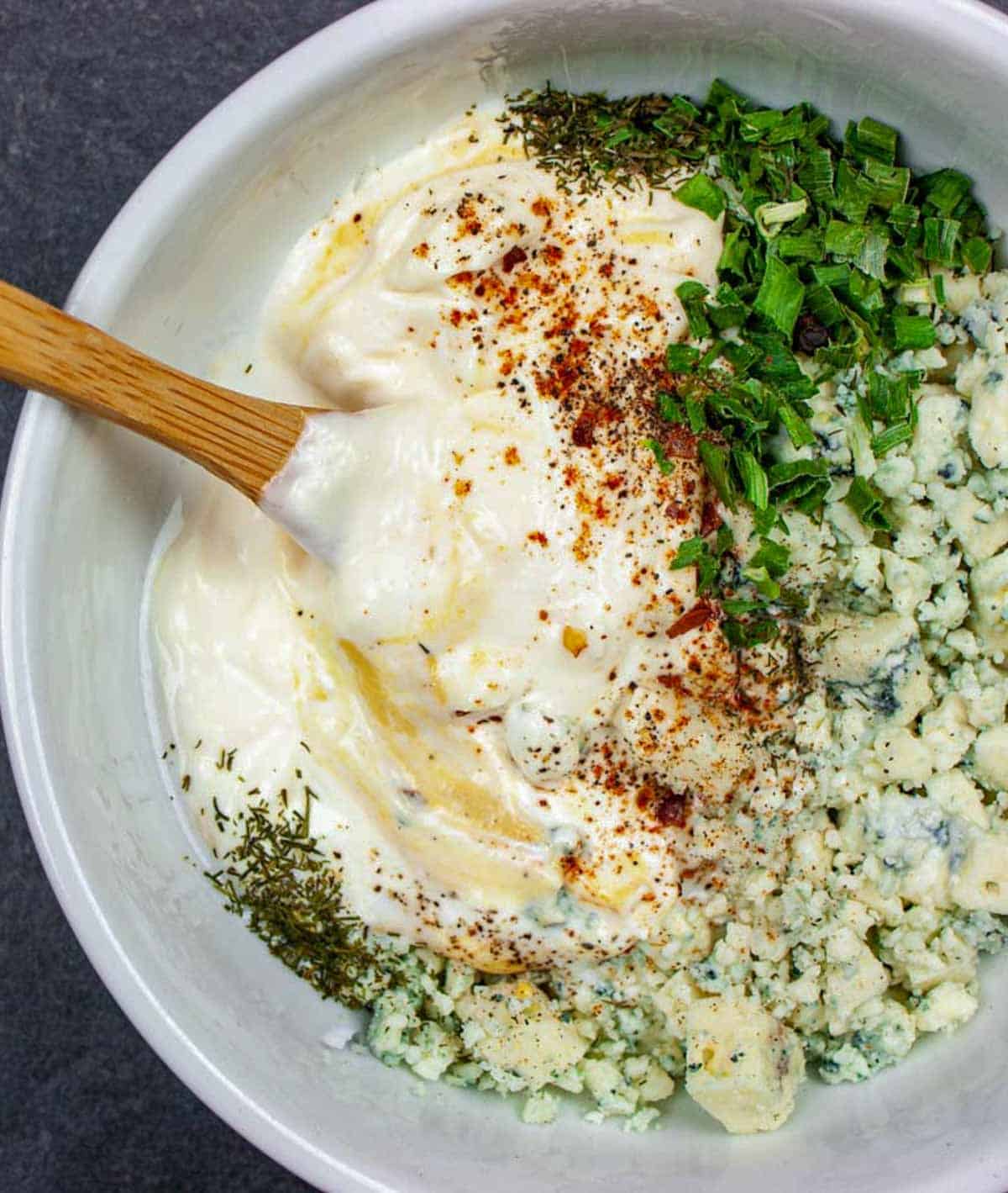 Unmixed blue cheese dressing ingredients in a white bowl with a small wooden spoon in it.