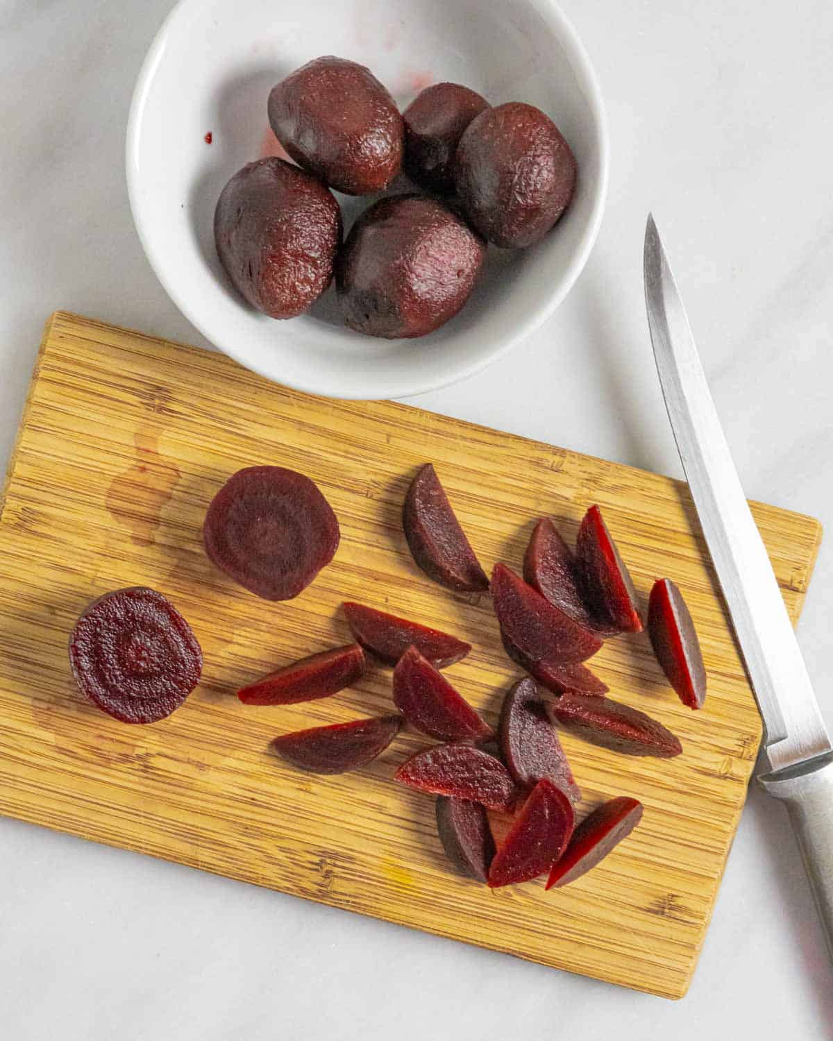 Cooked red beets in a small white bowl and on a board sliced into wedges.