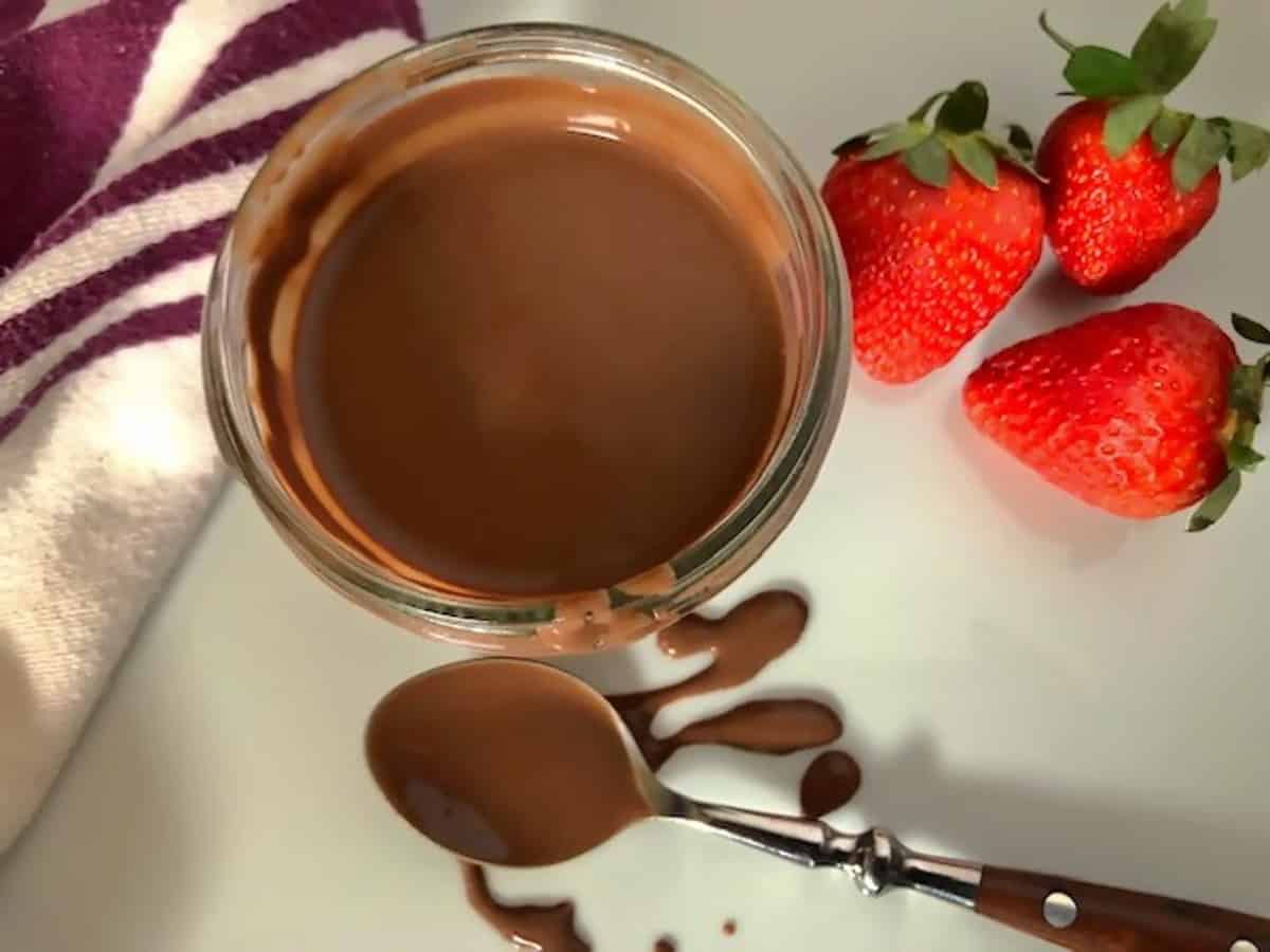 Keto chocolate sauce in a small mason jar with a spoon strawberries and a towel.