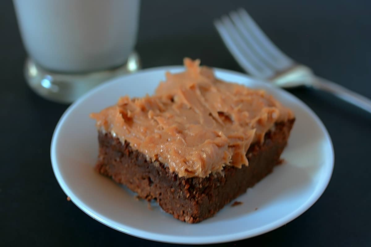 Keto chocolate brownie with peanut buttercream frosting on a white plate with a fork and glass of almond milk.