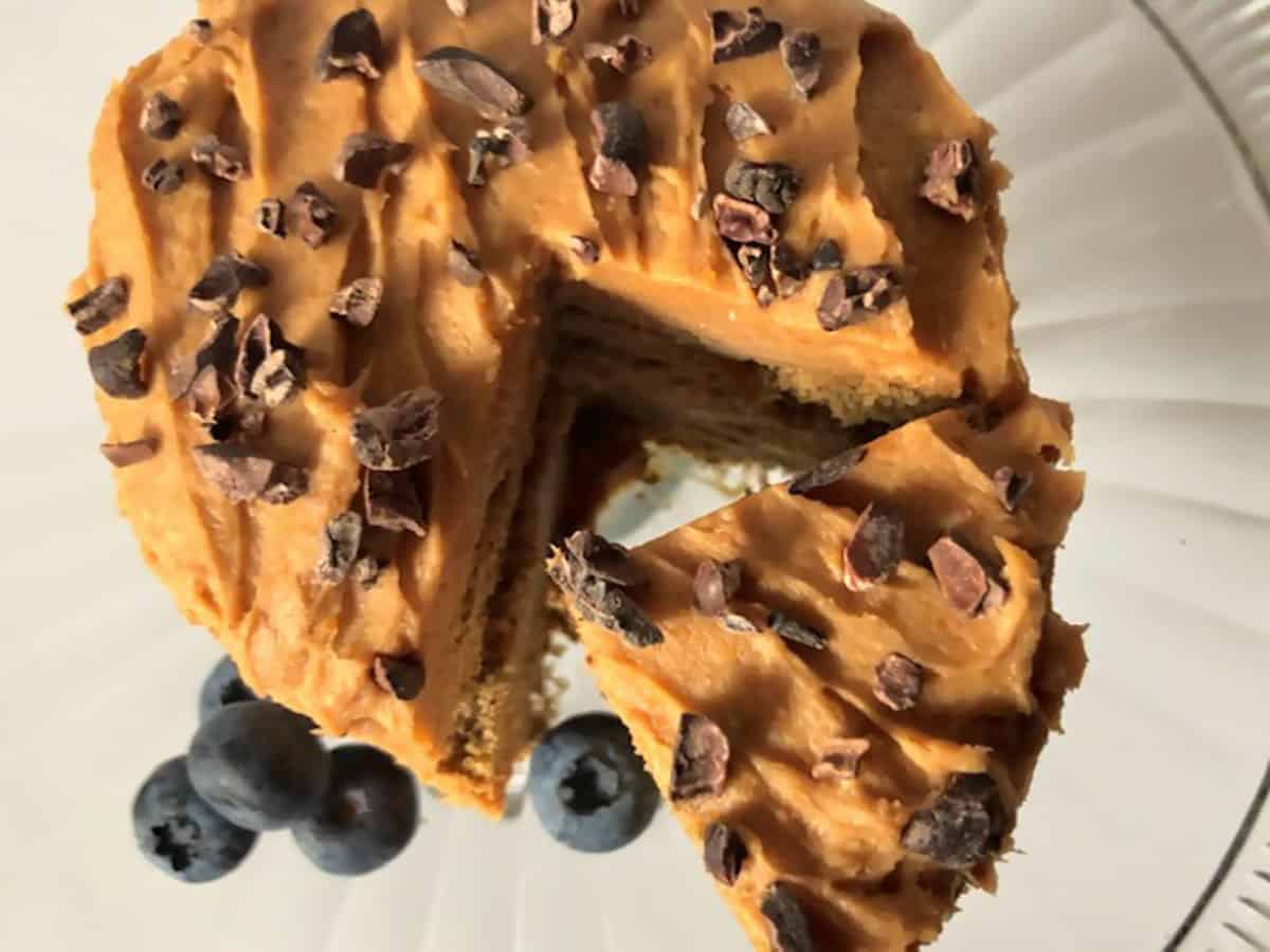 Cookie cake topped with peanut butter cream cheeze frosting and cacao nibs on a glass plate with blueberries on the side.