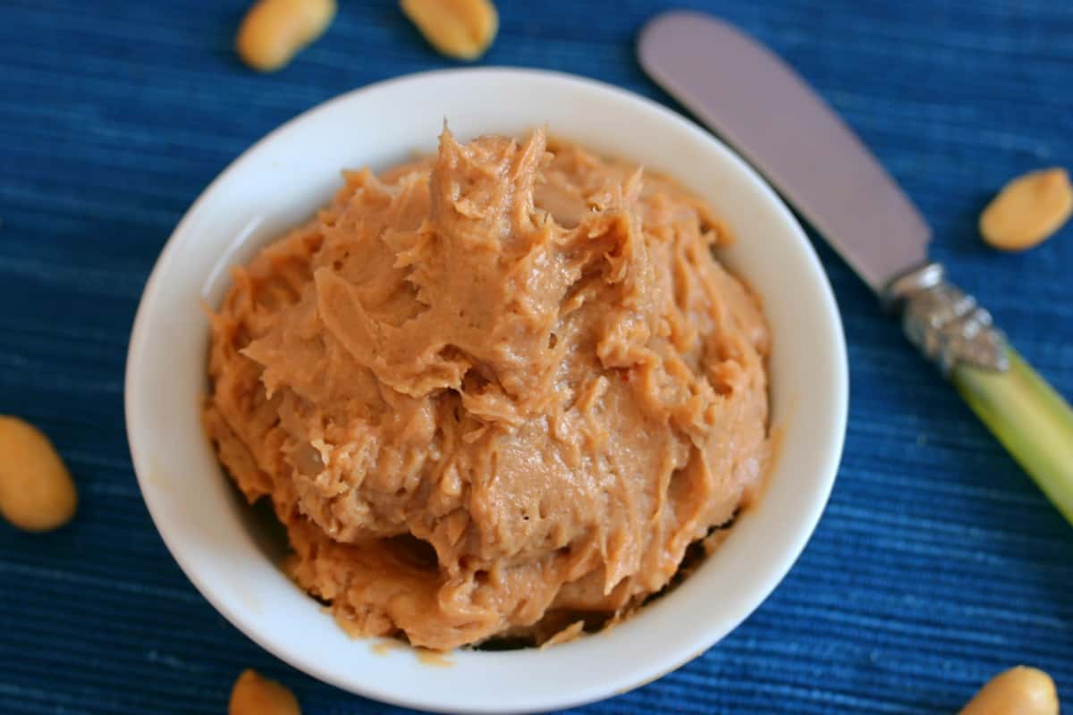 Peanut butter cream cheese frosting in a white bowl with a knife on a blue background with peanuts.