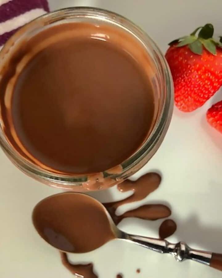 Dish of keto chocolate sauce with a dipped spoon on the side and three fresh strawberries.