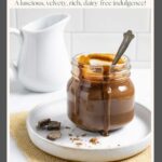 Small glass jar of sugar-free chocolate sauce with a spoon in it and sauce dripping down the sides, on a plate with cacao powder and chocolate chunks.