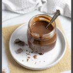 Glass jar of keto chocolate syrup with a spoon in it and sauce dripping down the sides, on a plate with cacao powder and chocolate chunks.