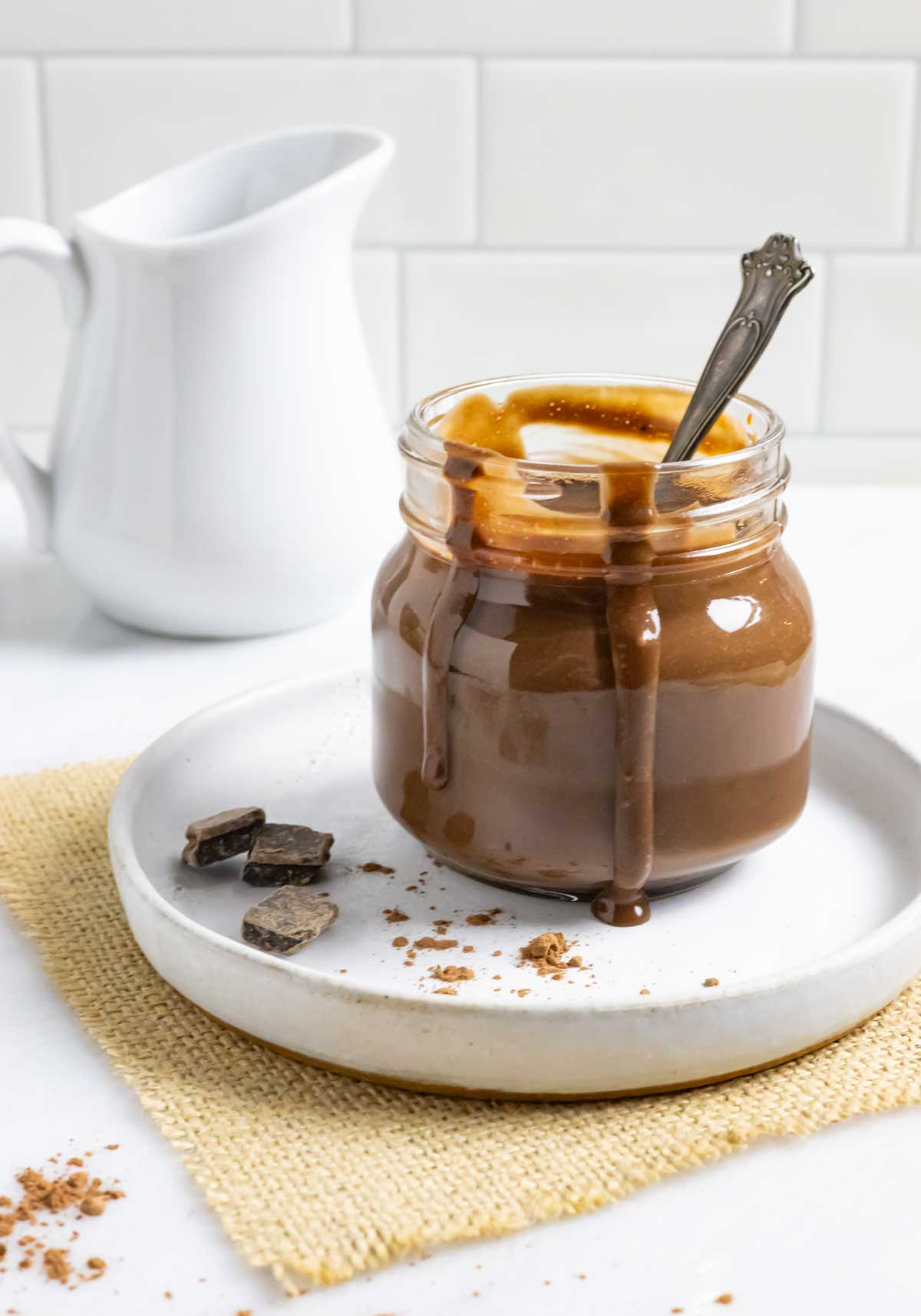 Small glass jar of sugar-free chocolate sauce with a spoon in it and sauce dripping down the sides, on a plate with cacao powder and chocolate chunks.