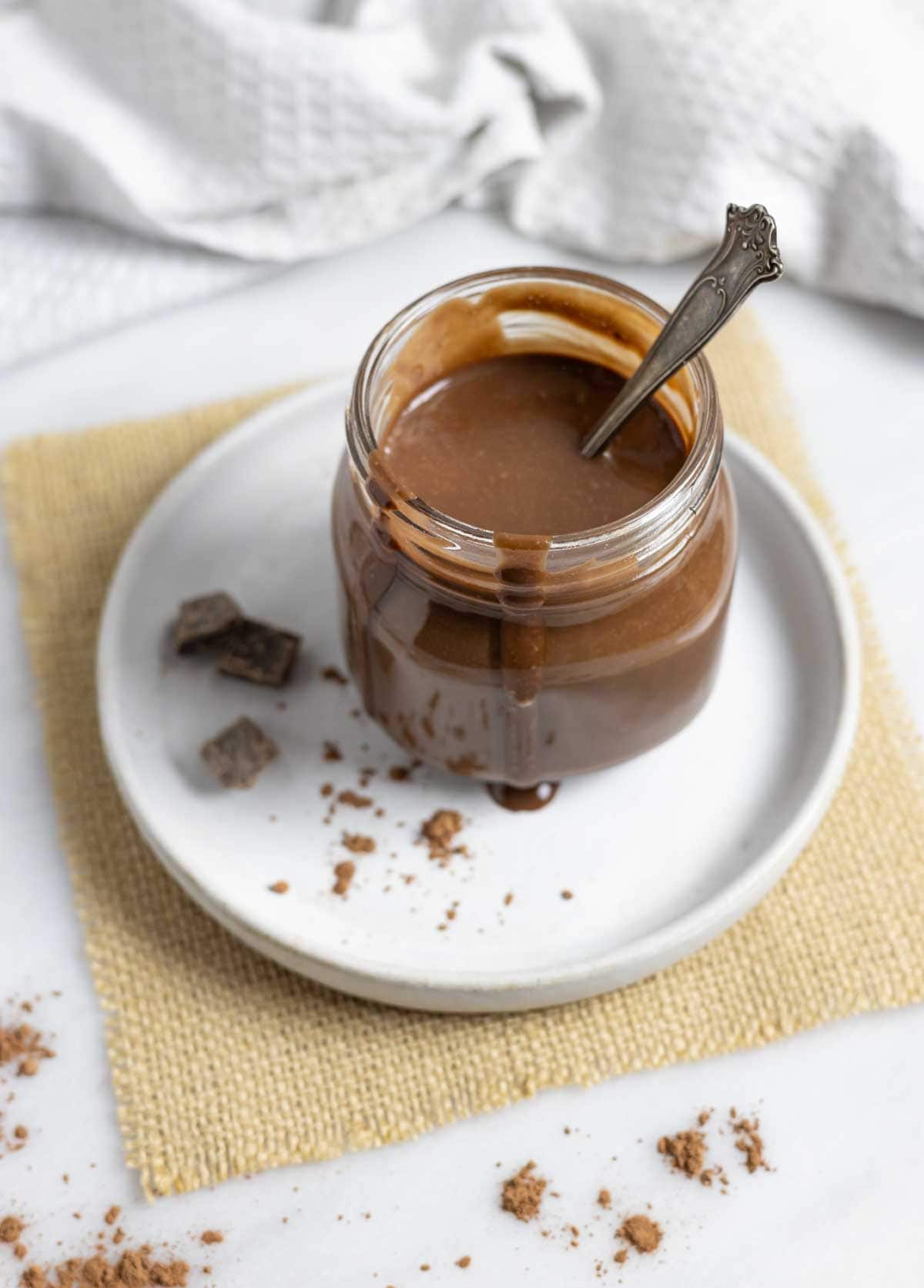 Glass jar of keto chocolate syrup with a spoon in it and sauce dripping down the sides, on a plate with cacao powder and chocolate chunks.