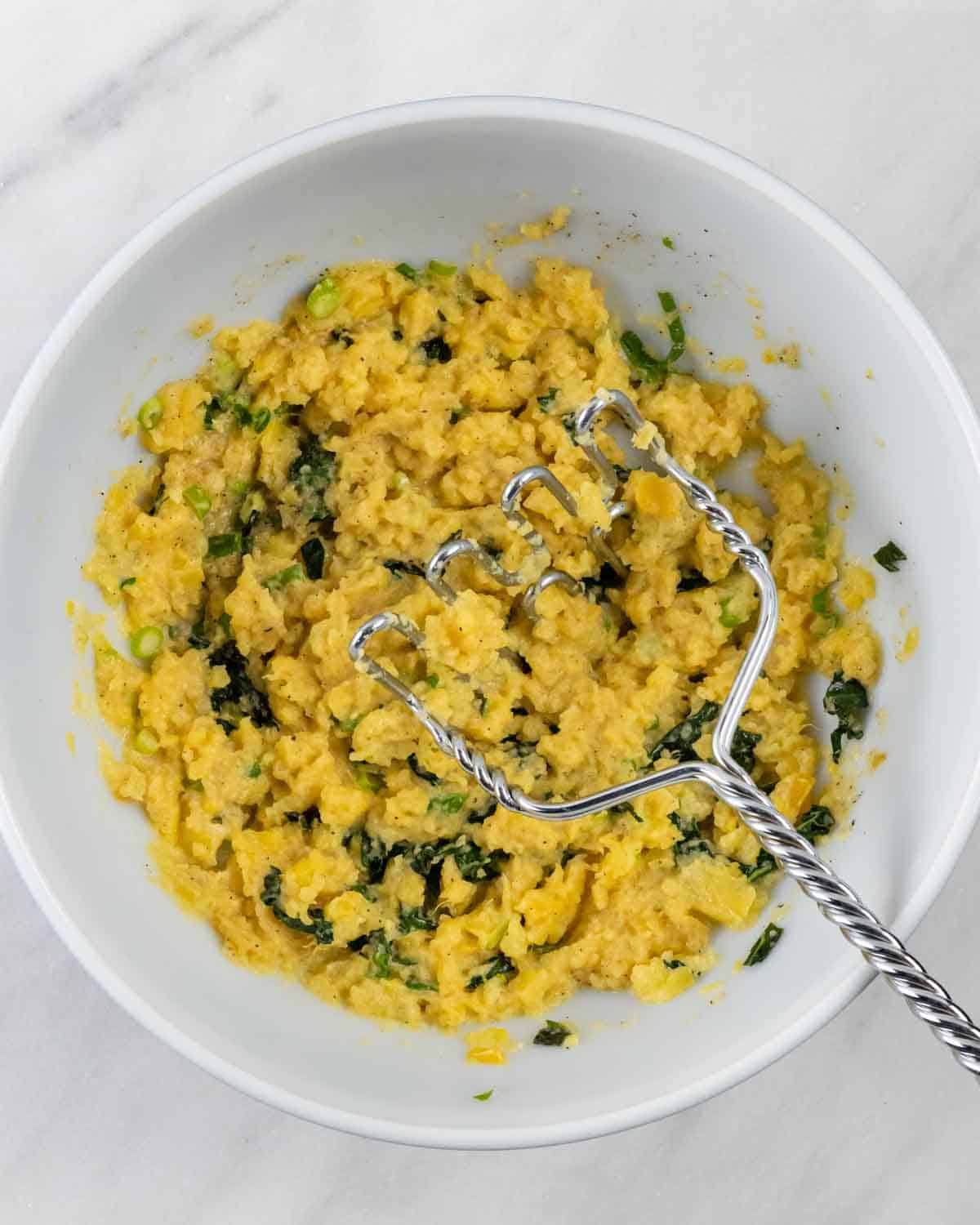 Mashed rutabagas with kale in a mixing bowl with a potato masher.