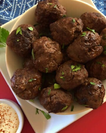 White serving bowl of keto corned beef meatballs on a white plate and red board, with ramekin of horseradish mustard sauce.