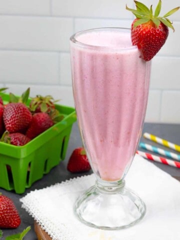 Tall glass of smoothie with a strawberry on the rim, sitting on a white napkin with colored striped straws and a green container of strawberries.