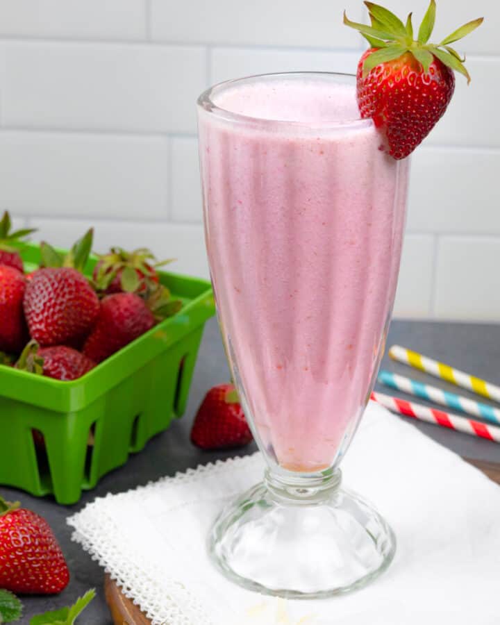 Tall glass of smoothie with a strawberry on the rim, sitting on a white napkin with colored striped straws and a green container of strawberries.