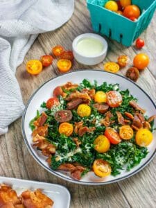 Kale blt salad with orange and red cut cherry tomatoes and bacon. Ramekin of dressing on the side with basket of tomatoes and plate of bacon.