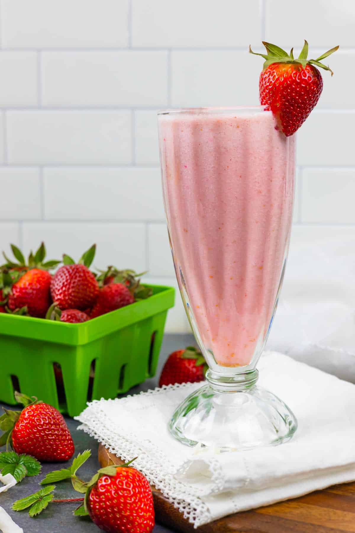 Shake glass of strawberry almond milk smoothie with a berry on the rim and a container of berries next to the glass.