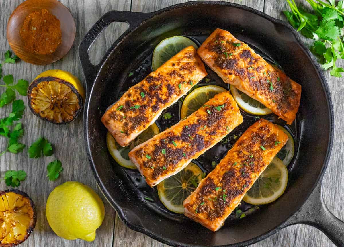 Four pan-fried Indian Salmon fillets on top of lemon slices in a cast iron skillet with seared lemon halves and a whole lemon next to it.