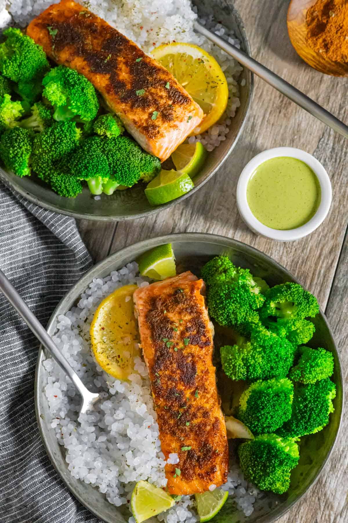 Two plates of Indian Salmon with broccoli, shirataki rice, lemon and lime wedges with a side of cilantro dressing.