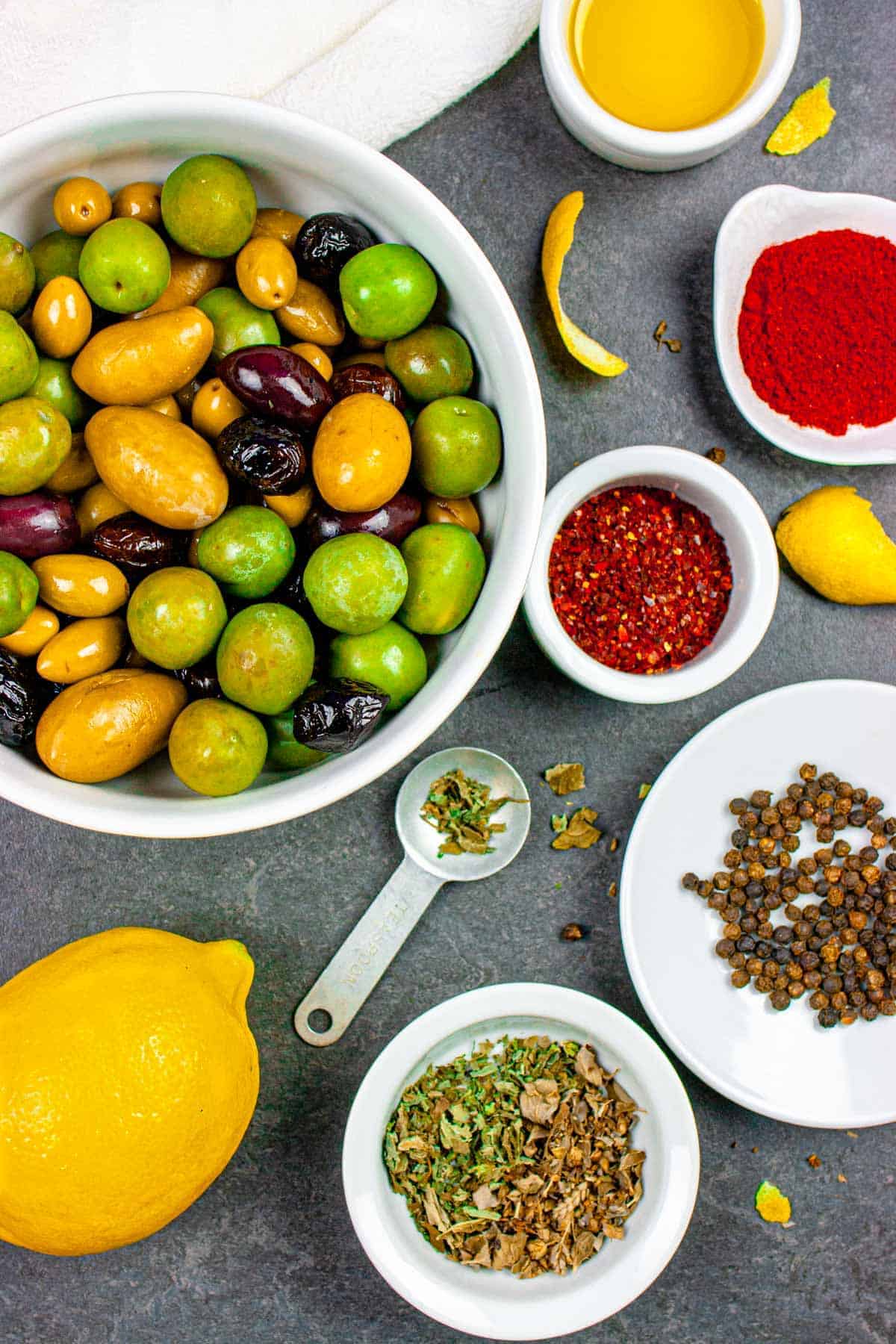 Ingredients for marinated olives including mixed olives in a bowl, spices in small ramekins, and a whole lemon on the side.
