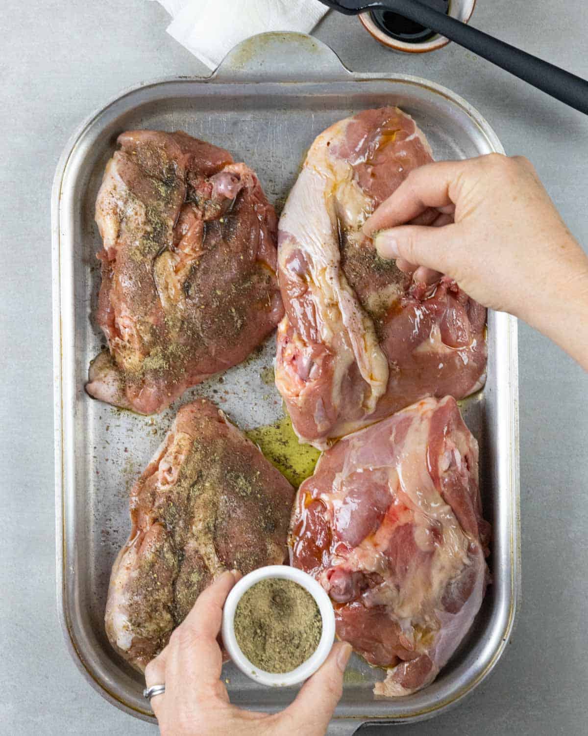 Sprinkling spice rub on underside of four turkey thighs in a steel pan.