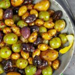 Spicy olives of many varieties in a serving bowl sprinkled with Aleppo pepper, on a grey board with a spoon and sprig of parsley.