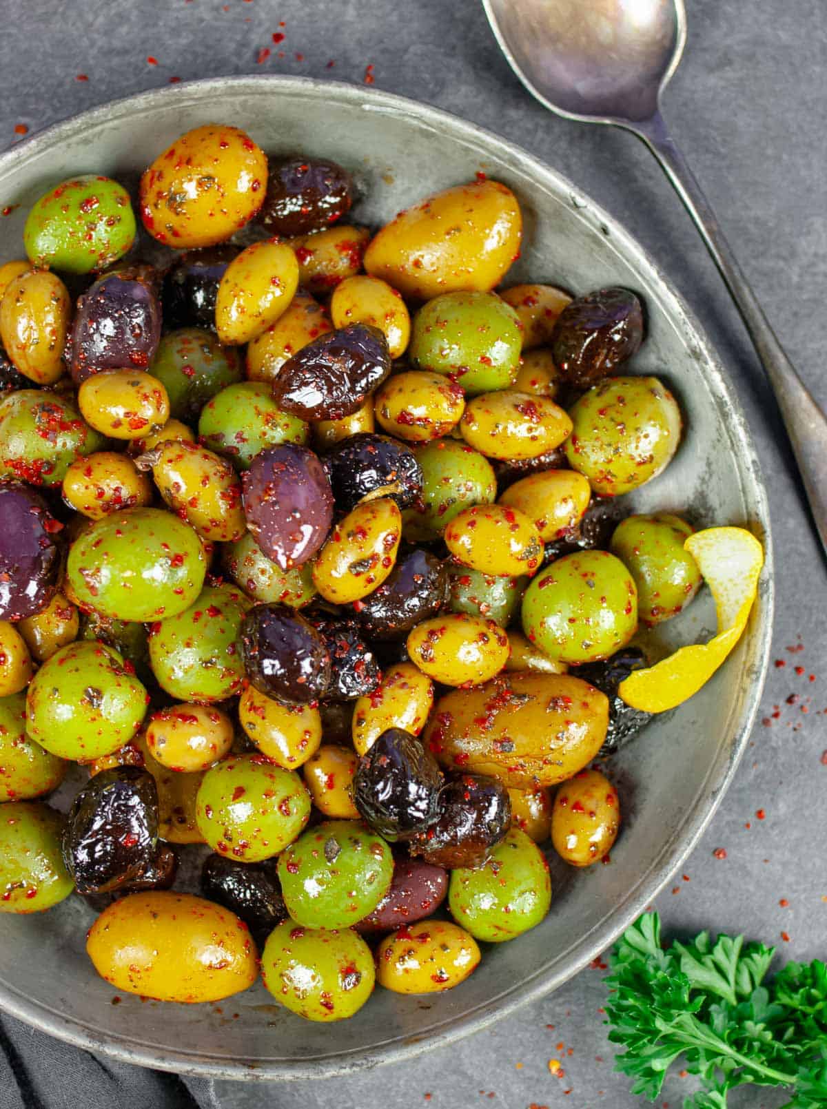 Spicy olives of many varieties in a serving bowl sprinkled with Aleppo pepper, on a grey board with a spoon and sprig of parsley.