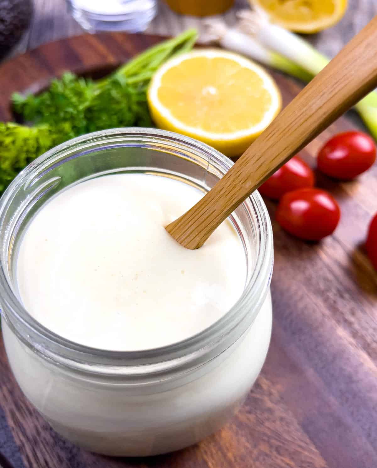 Small mason jar of lemon dressing with a wooden spoon in it, on a board with garnish of parsley, cherry tomato, and a cut in half lemon.