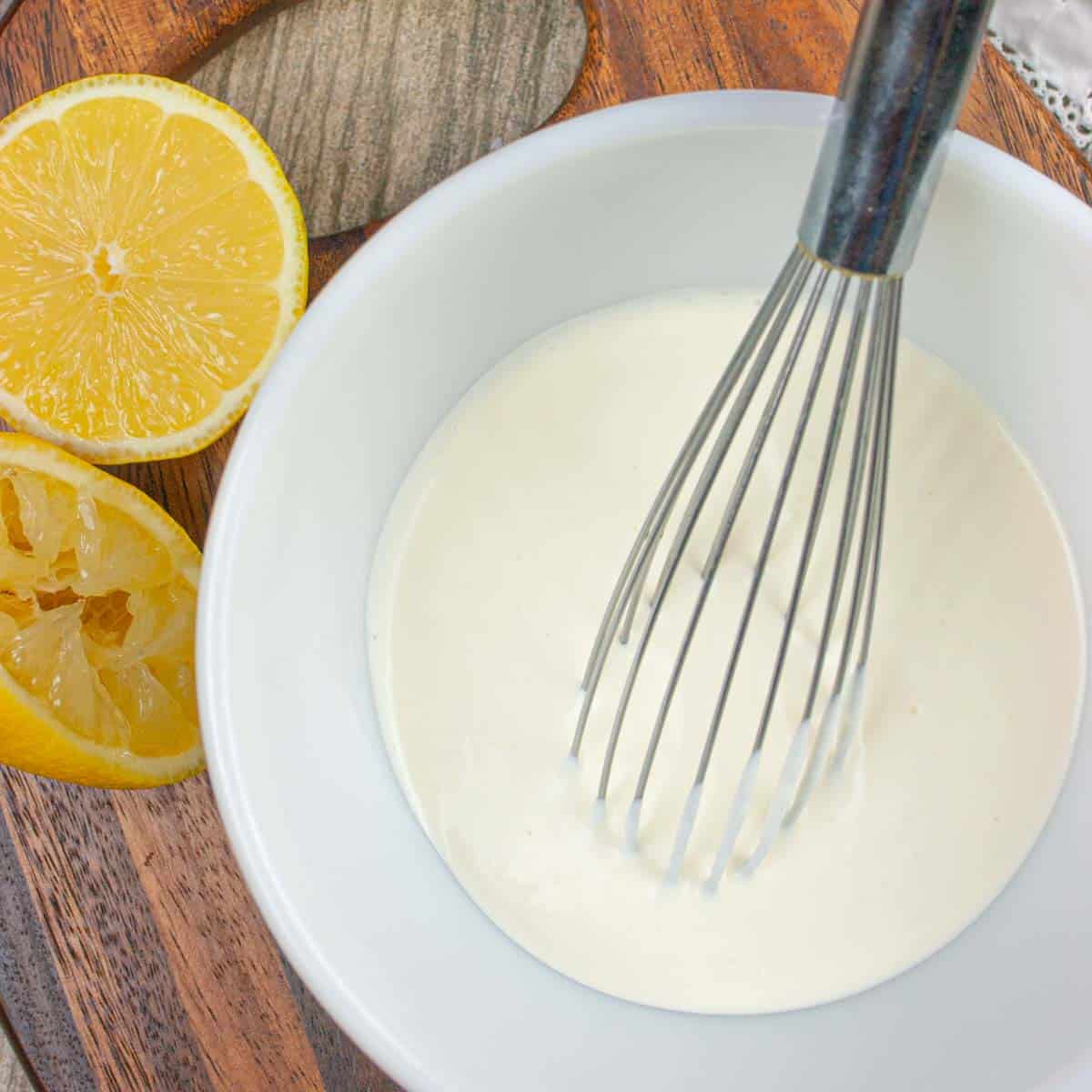 White mixing bowl containing creme fraiche dressing and a whisk, sitting on a board with cut lemons and a wooden spoon.
