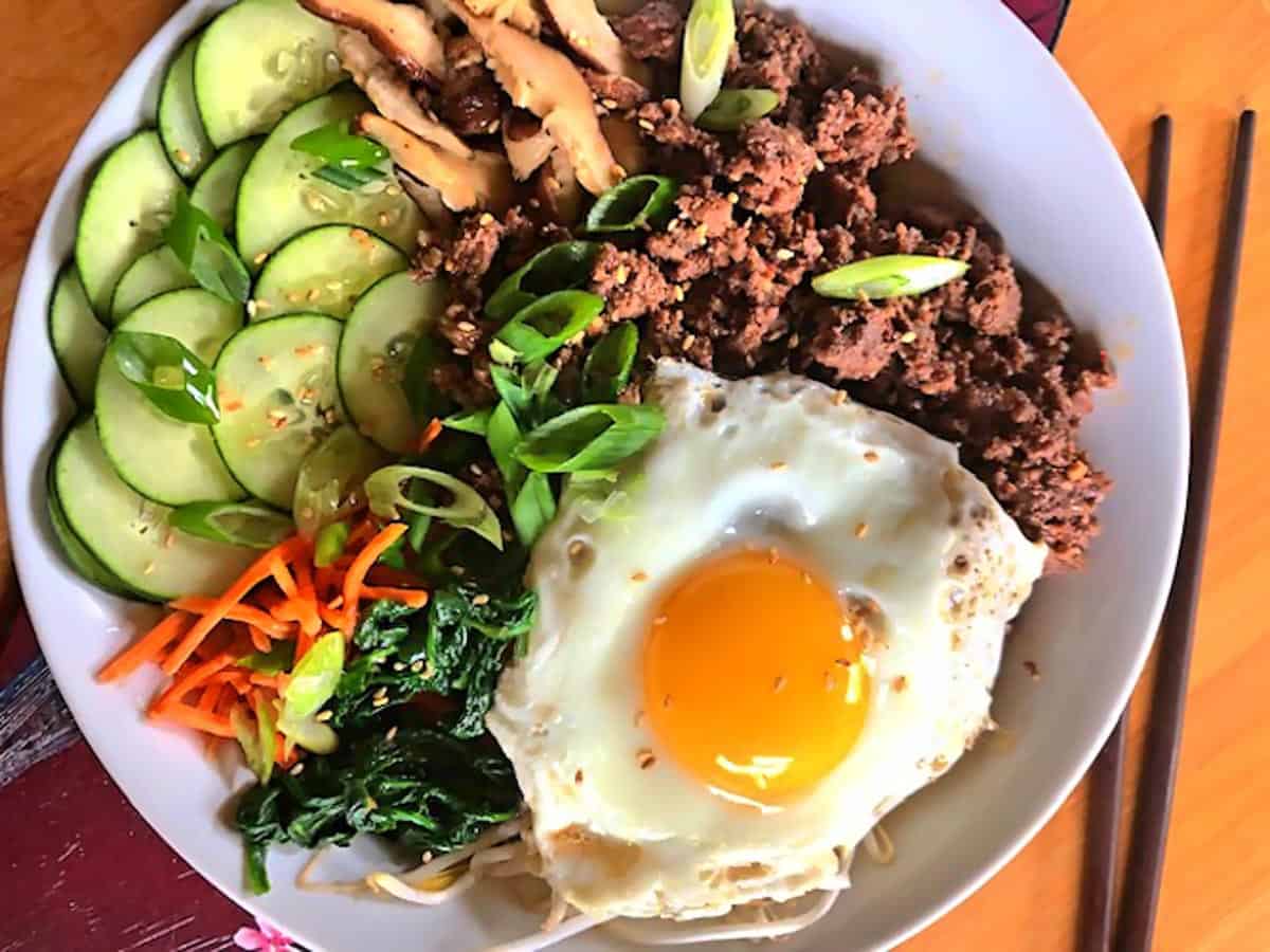 Bibimbap in a white bowl on a painted board a sunnyside up egg on top and chopsticks on the side.