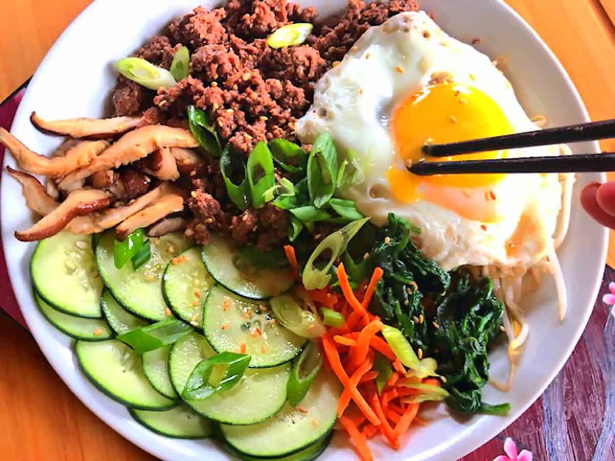 Bibimbap in a white bowl on a painted board with chopsticks popping a sunnyside up egg yolk on top.